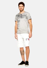Tricou grafic gri cu text variat; pantaloni scurți albi până la genunchi cu imprimeu roșu; adidași negri; tatuaj vizibil pe piciorul stâng.
