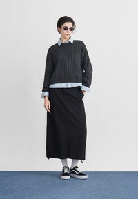 Άτομο που φοράει μαύρο πουλόβερ πλεκτό με καλώδια πάνω από μπλε πουκάμισο, μακριά μαύρη φούστα, λευκές κάλτσες, μαύρα αθλητικά παπούτσια και στρογγυλά γυαλιά ηλίου, στέκεται σε μπλε πάτωμα.