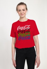 Červené bavlněné tričko s opakujícím se textem "Coca-Cola" v bílé, černé, zelené a modré barvě, s volným střihem a krátkými rukávy.