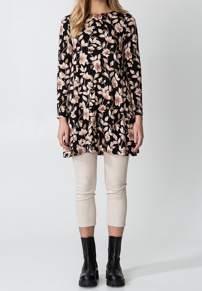 Tunique noire à motif floral, manches longues, ornée de fleurs roses et crème. Associée à un pantalon beige raccourci et des bottines noires.