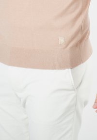 Pull en maille beige clair avec bord-côtes en bas et aux poignets, présentant un petit logo brodé sur le côté gauche. Assorti avec un pantalon blanc.