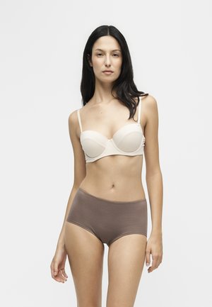 Kvinde med langt mørkt hår iført en lysebeige bh og brune højtrissede trusser, stående mod en ensfarvet lys baggrund.