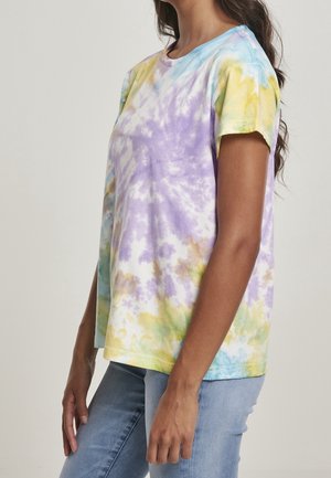 Kvinde iført pastel tie-dye kortærmet T-shirt i lilla, gul, blå og grøn med lyseblå jeans, vist fra siden.