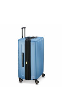 Maleta rígida en azul claro, con textura acanalada vertical, cremallera negra, ruedas dobles y un asa extensible para un transporte fácil.