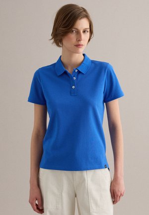 Junge Frau mit kurzen braunen Haaren, die ein blaues Polohemd und helle Hosen trägt, steht vor einem schlichten Hintergrund.