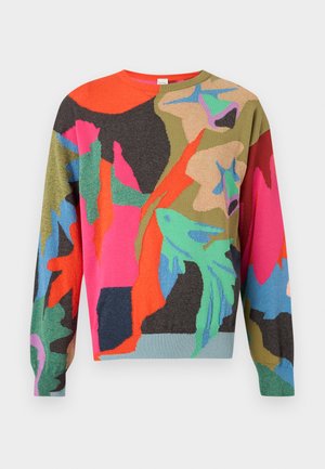 Muž nosiaci viacfarebný abstraktný vzorovaný sveter, béžové nohavice s manžetami, čierne mokasíny a modrú košeľu s golierom pod ním.