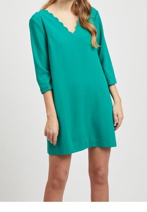 Žena v tyrkysově modrých šatech ke kolenům s vroubkovaným výstřihem do V a tříčtvrtečními rukávy, stojící před jednobarevným pozadím.