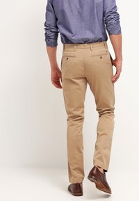 Chino beige in cotone con vestibilità slim. Presenta due tasche posteriori e una finitura liscia. Indossato con una camicia blue a maniche lunghe e scarpe marroni.