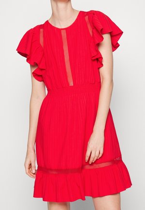 Frau trägt ein hellrotes Kleid mit Rüschen an Ärmeln und Saum, mit transparenten vertikalen Streifen am Oberteil und nahe am Rocksaum.