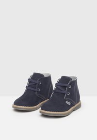 Ein Paar navyblaue Wildleder-Kinderstiefel mit beigen Sohlen und zwei Ösen zum Schnüren auf einer weißen Oberfläche.