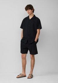 Chemise noire à boutons, à manches courtes, en tissu texturé ; associée à un short noir assorti. Sandales beiges à une seule lanière.