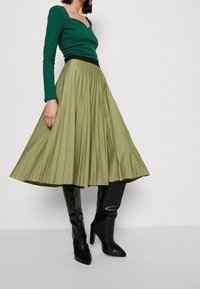 Top verde de manga larga con canalé combinado con una falda midi plisada verde oliva y botas negras de tacón alto hasta la rodilla. Diseño limpio con tejido texturizado.
