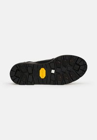 Suela de goma negra de un zapato con surcos profundos, manchas naranjas y un logo amarillo de Vibram en el centro sobre un fondo blanco.