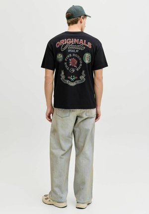 Moški, oblečen v črno majico z grafiko z rdečim in zelenim besedilom, svetlo sive kavbojke, bež superge in zeleno baseball kapo, obrnjeni stran na belem ozadju.