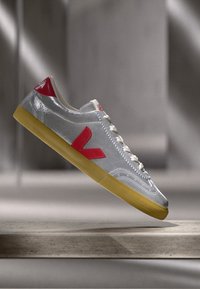 Gray synthetic sneakers with a shiny finish, red accents, and a textured yellow rubber sole. Features a lace-up design and logo detailing.