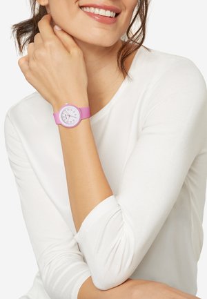Pink silikoneur med en rund hvid urskive, markerede time- og minutvisere og pink timemarkører. Båret på et håndled iført en hvid skjorte.