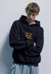 Černá mikina s kapucí s nápisem "Jedno poslední zvláštní dobrodružství" žlutým písmem. Měkká tkanina, prostorný střih a žebrované manžety. Spárováno se šedými skládacími kalhotami.