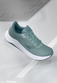 Zaļas Under Armour skriešanas kurpes ar baltu zoli, ar acs virsu un mīkstinātu apkakli, parādītas pret pelēku iekštelpu sporta grīdas fona.