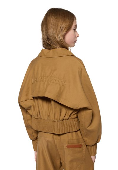 Mädchen mit hellbraunen Haaren, das eine braune kurze Jacke mit "LANVIN" auf dem Rücken und passende Hosen trägt, steht seitlich.