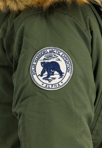 Grønn parka med hette fôret med pels, med et sirkulært brodert merke med en blå bjørn og teksten "Polar Warriors Arctic Expedition".
