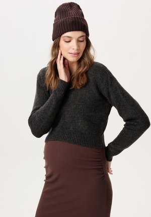 Zwangere vrouw die een bruine muts, een donkergrijze trui en een bruine rok draagt, zacht haar gezicht aanrakend en zacht glimlachend.