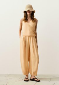 Femme portant un pantalon beige à jambes larges et un top cache-cœur sans manches avec un grand chapeau de soleil beige tissé et des sandales à plateforme noires.
