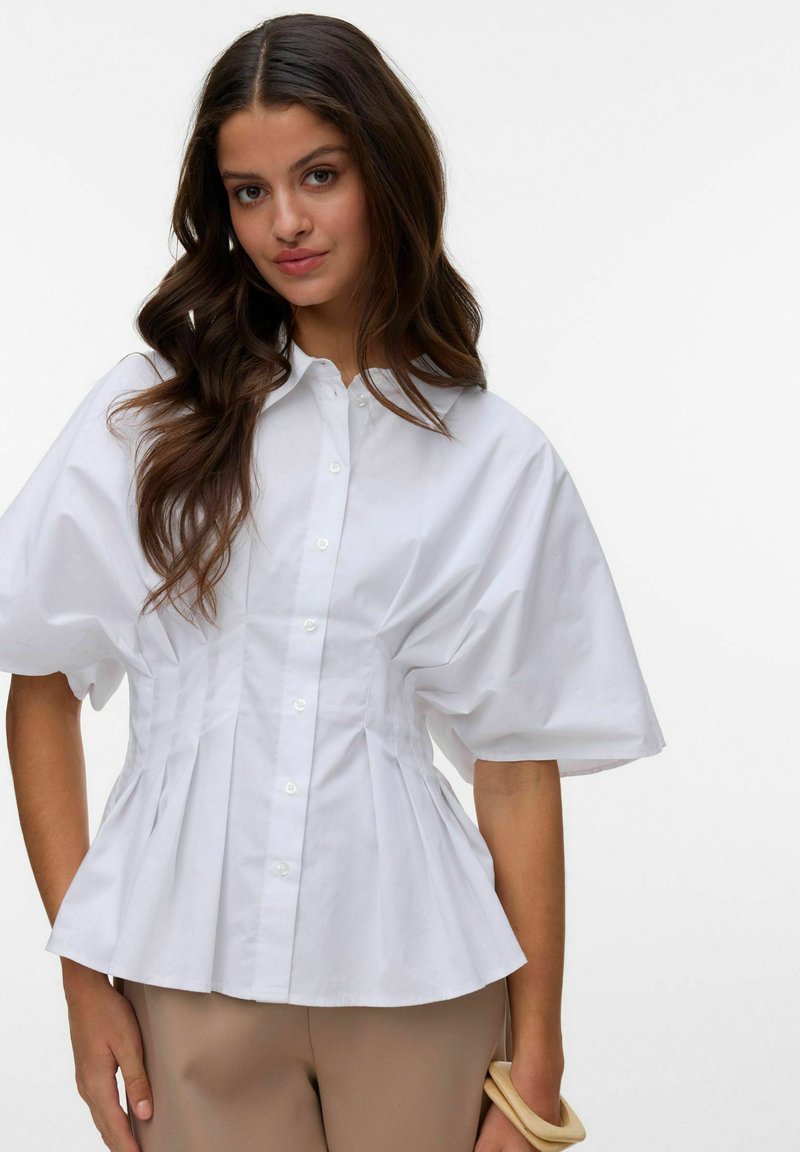 Femeie cu păr lung și castaniu poartă o cămașă albă cu mâneci bufante și nasturi și pantaloni bej, ținând un brățară de lemn pe încheietura mâinii.