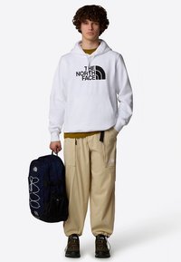 Biela mikina s kapucňou a čiernym logom, béžové cargo nohavice a námornícky modrý batoh s vertikálnymi vreckami a čiernymi detailmi.