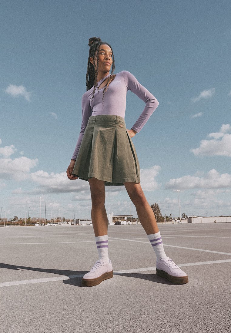 Haut lilas à manches longues, jupe plissée olive, chaussettes blanches avec des rayures violettes, et chaussures blanches avec des semelles brunes, sur fond de ciel bleu.