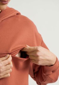 Hoodie in Rostfarbe mit Reißverschlusstasche. Aus Baumwolle, lockere Passform, gerippte Manschetten. Hand zieht den Reißverschluss auf, um den inneren Stoff zu zeigen.