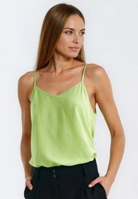 Zielona satynowa koszulka na cienkich ramiączkach, z dekoltem w kształcie litery V i luźnym krojem, zestawiona z czarnymi, wysokim stanem spodniami. Gładka faktura i jasny kolor.