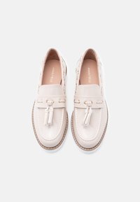 Mocassins en cuir verni beige avec lacets décoratifs, accents de pompons et coutures contrastées sur une semelle de couleur claire. Design flexible et léger.