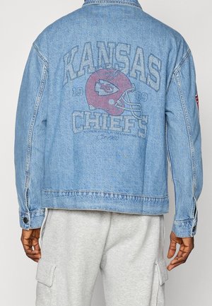 Tył jasnoniebieskiej kurtki dżinsowej z wyblakłym czerwonym logo kasku futbolowego Kansas Chiefs i napisem, noszonej przez osobę w szarych spodniach typu cargo.