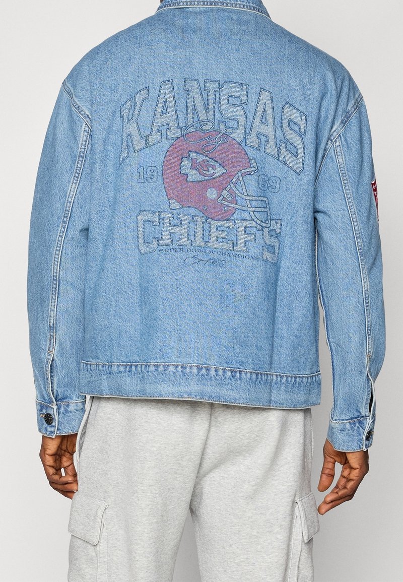 Dos d'une veste en jean bleu clair avec un logo et un texte rouge délavés du casque de football américain des Kansas Chiefs, portée par une personne en pantalon cargo gris.
