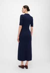 Robe en maille côtelée marine avec col polo, manches courtes, silhouette ajustée et longueur maxi, associée à des chaussures bourgogne brillantes.
