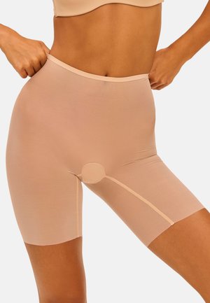 Frau justiert hoch taillierte, hautfarbene Shaping-Shorts, die bis zur Mitte des Oberschenkels reichen, gezeigt vom mittleren Oberkörper bis zu den Oberschenkeln vor einem einfarbigen Hintergrund.