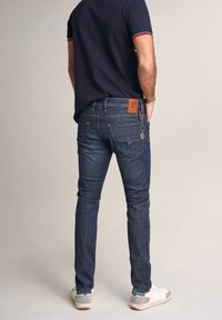Jeans de mezclilla azul oscuro con un diseño ajustado, bolsillos traseros y un parche de cuero. Combinados con una polo de manga corta en color navy y zapatillas deportivas.