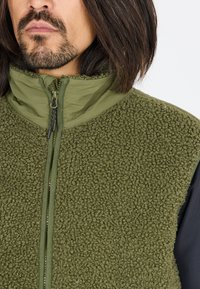 Groene fleece vest met een hoge kraag, textuurstof, volledige ritssluiting en gelaagde ontwerpdetails. Geaccentueerd met een gladde stof eronder.