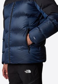 Granatowa kurtka puchowa z pikowanym wzorem, z zamkiem błyskawicznym z przodu i widoczną kieszenią boczną. Na mankiecie znajduje się etykieta "700".