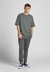 Graues oversized T-Shirt mit kurzen Ärmeln und dezentem Logo; kombiniert mit dunkelgrauen Trainingshosen; Model trägt schwarze Sneakers mit weißen Sohlen.