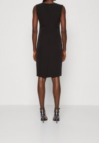 Schwarzes ärmelloses Kleid mit einem subtilen Schlitz auf der Rückseite. Gefertigt aus glattem Stoff und mit einer taillierten Silhouette. Ergänzt durch glänzende graue High Heels.