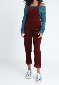 Kvinna klädd i vinröda kordoveraller, en turkos avknäppt tröja och flerfärgad sneakers, står mot en helt vit bakgrund.