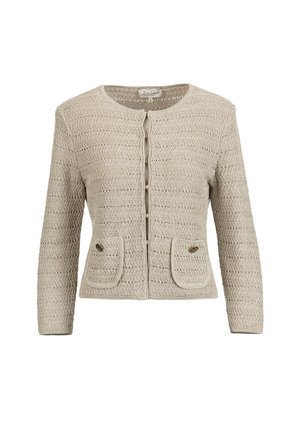 Frau trägt beigefarbene, strukturierte Jacke mit Knöpfen und passenden knielangen Rock, hält eine cremefarbene Handtasche mit goldener Kette, steht vor weißem Hintergrund.