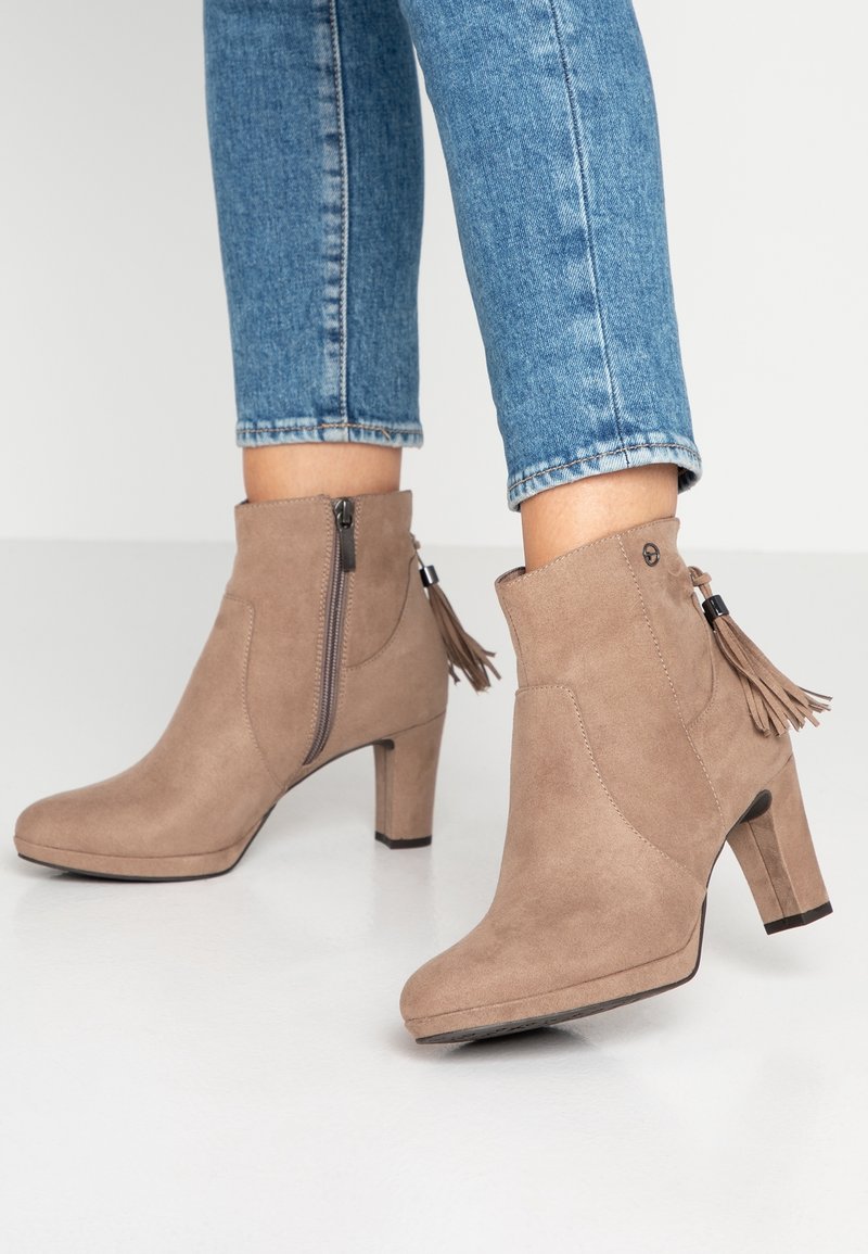 Bottines en daim beige à talons carrés, zips latéraux, détails de pompons, portées avec un jean denim bleu coupé au-dessus de la cheville, sur un sol clair.