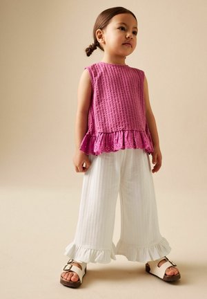 Junges Mädchen mit braunen Haaren, die zu einem tiefen Dutt gebunden sind, trägt ein ärmelloses rosa Oberteil mit Rüschensaum, weiße, weit geschnittene Hosen mit gerüschten Bündchen und weiße Sandalen.