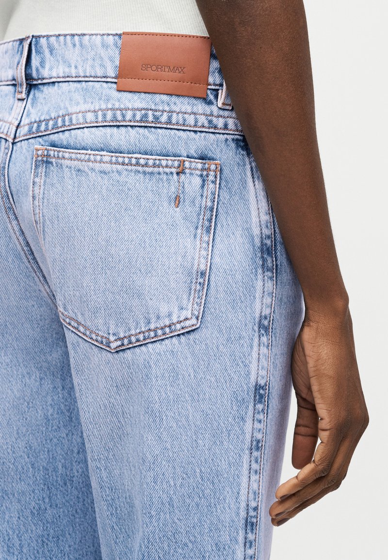 Close-up van een persoon die lichtblauwe spijkerbroek draagt met een leren label met de tekst "SPORTMAX" nabij de tailleband.