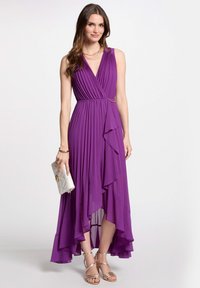 Robe plissée violette avec un col en V et un ourlet asymétrique, accentuée par une chaîne dorée à la taille. Portée avec une pochette argentée et des talons dorés.