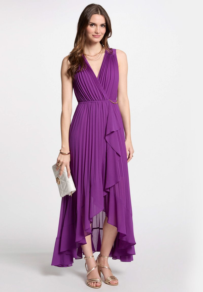 Robe plissée violette avec un col en V et un ourlet asymétrique, accentuée par une chaîne dorée à la taille. Portée avec une pochette argentée et des talons dorés.