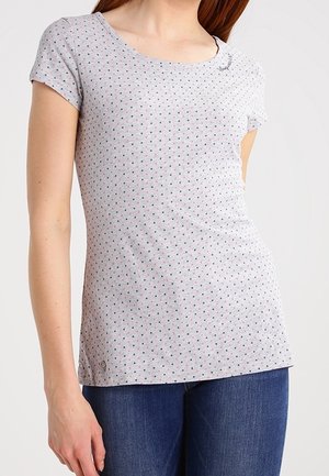 Žena v upnutém světle šedém tričku s krátkým rukávem, pokrytém drobnými růžovými a černými puntíky, a modrých džínách.