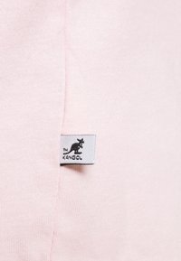Jasnoróżowa tkanina o miękkiej teksturze, z białą metką z czarnym logo kangura oraz napisem "KANGOL" blisko szwu.